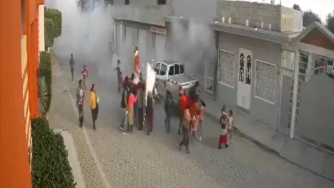 Una vista aérea muestra una concurrida calle de la ciudad con personas y vehículos. En el lado izquierdo, un edificio alto se eleva sobre el resto, dominando el horizonte. En medio del humo blanco tras estallido de pirotecnia.