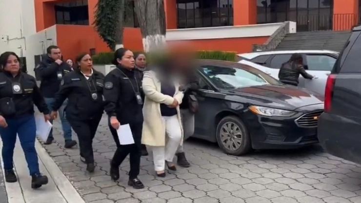 Una mujer de mediana edad, con el cabello recogido y vestida con un traje oscuro, está sentada en un gran sillón de madera tallada con reposabrazos, detrás de un escritorio oscuro. Su rostro está cubierto por una barra negra horizontal para preservar su anonimato. A su izquierda, sobre el escritorio, hay una pequeña bandera de México. El fondo es una pared con azulejos texturizados en tonos claros.