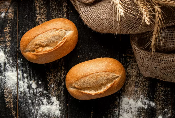 Dos panes tipo bolillo sobre una mesa de tabla con un poco de harina y trigo.