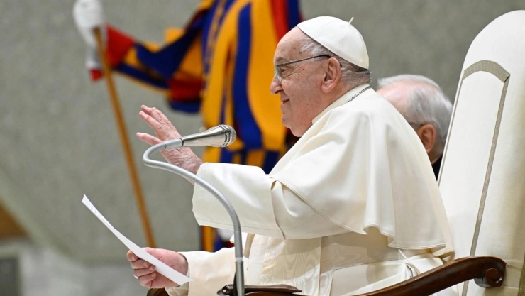Papa Francisco, sentado, frente a un micrófono, con una hoja de papel en la mano derecha, y la mano izquierda levantada.