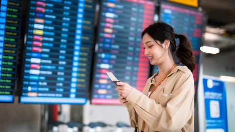 Una joven con el pasaporte en la mano delante de las pantallas que indican los vuelos en un aeropuerto