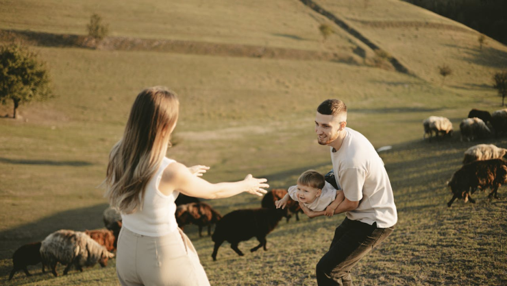 Mujer con los brazos abiertos, juega con su bebé y su esposo vestido de blanco en el campo.