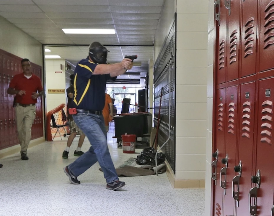 Un profesor del distrito escolar de Clarksville, con máscara protectora en el fondo, usa una pistola de práctica mientras se dirige a un aula en la la secundaria básica de Clarksville, Arkansas, mientras estudiantes hacen el papel de víctimas en un simulacro de tiroteo