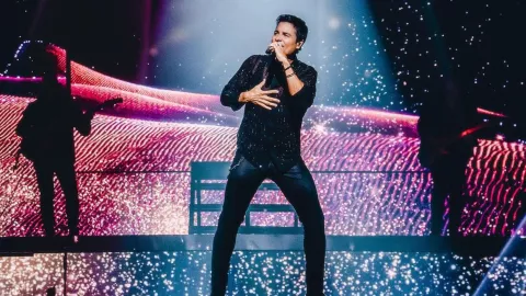 Chayanne con un micrófono cantando en su tour “bailemos otra vez”, vestido de negro con una camisa con brillos