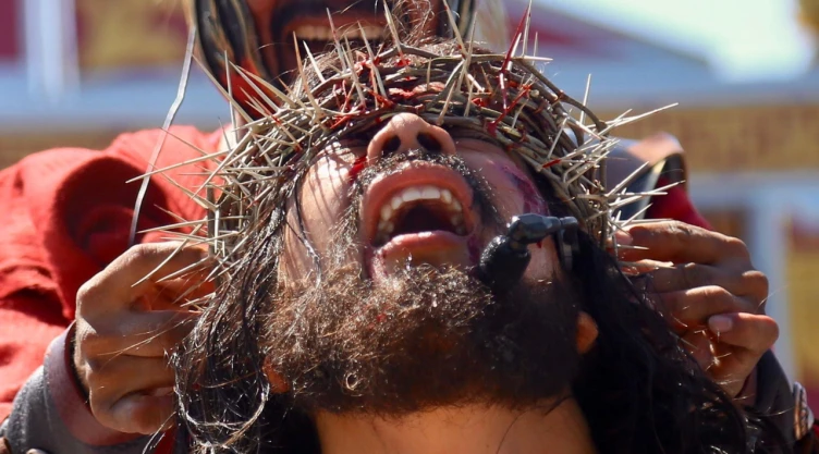 Primer plano dramático de Arnulfo Morales interpretando a Jesús en la Pasión de Cristo de Iztapalapa 2026. Se observa al actor con una corona de espinas real sobre la frente, rastros de sangre en el rostro y expresión de dolor intenso con la boca abierta. En el fondo, se ven las manos de un soldado romano interpretado por otro actor y que ajustando la corona.