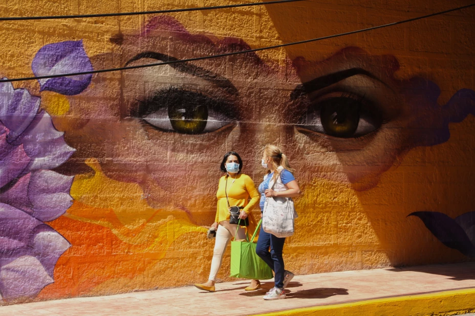 Dos mujeres caminan frente a un mural de grandes ojos, intervenido por artistas urbanos.