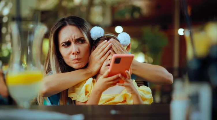 Una mujer cubre los ojos de una niña que tiene un teléfono en la mano.