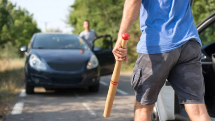 Un hombre con una camiseta azul y pantalones cortos de carga se baja de un coche blanco, sosteniendo un bate de béisbol con la punta roja. Detrás de él, en la distancia, otro hombre se asoma desde la puerta abierta de un coche oscuro estacionado en la carretera. El fondo es un camino rural con árboles y vegetación borrosa bajo la luz del día.