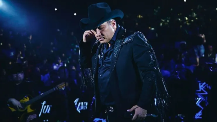 Julión Álvarez vestido de negro con un sobrero de vaquero agarrándose la oreja con cara de preocupación