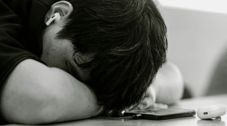 Foto de adolescente recostado sobre mesa con teléfono celular.