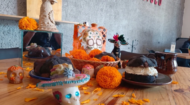 Mesa de madera decorada con piezas de pan de muerto negro, una rellena de crema chantilly blanca y otra con cubierta de chocolate, rodeadas de flores de cempasúchil, calaveritas de azúcar y una taza de barro.