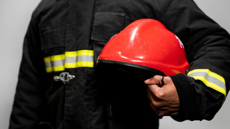 Bombero con uniforme negro y franjas reflejantes sostiene un casco rojo de protección en la mano.