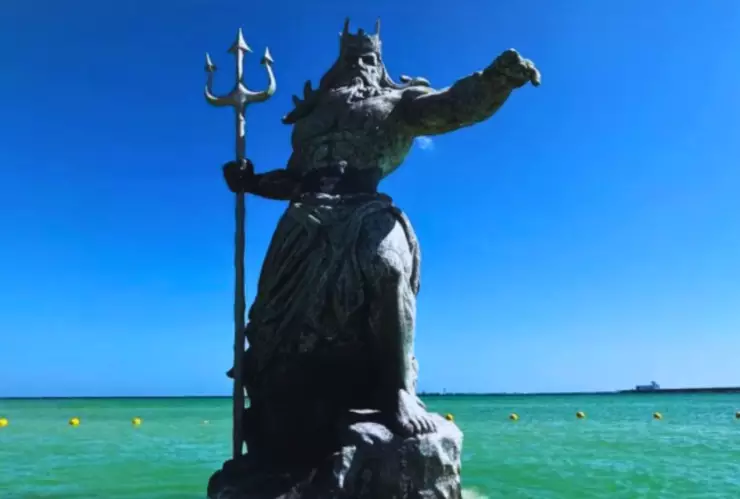 Es un hombre de barba larga apuntando hacía el lado derecho con un tridente en la mano y una corona sobre su cabeza