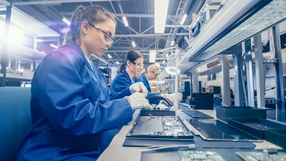 Woman Electronics Factory Worker in Blue Work Coat and Protective Glasses is Assembling Laptop’s Motherboard with a Screwdriver. High Tech Factory Facility with Multiple Employees.