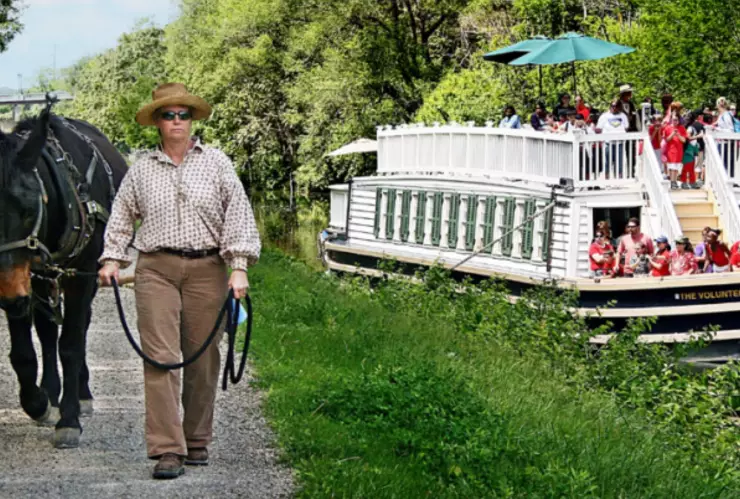 Hombre con un caballo de tiro caminando por el sendero a la vera del río, y una pequeña embarcación con turistas paseando por la zona.