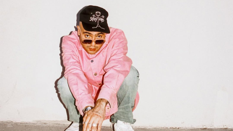 Milkman, rapero mexicano, posa en cuclillas con camisa rosa, gorra negra y gafas oscuras frente a un fondo blanco.