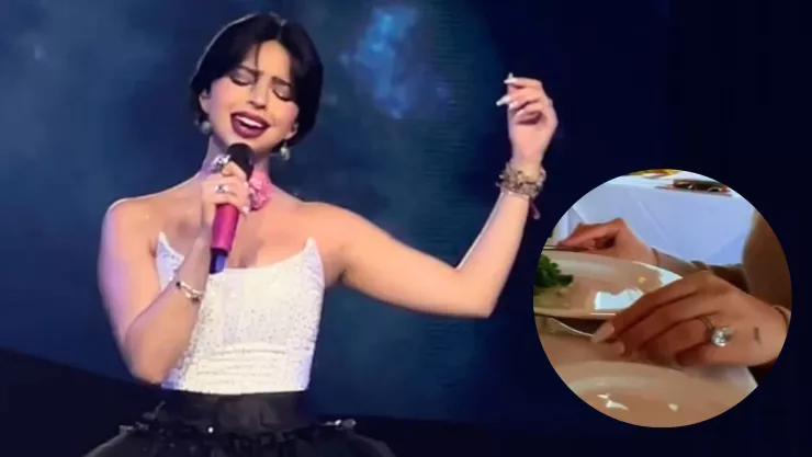 Ángela Aguilar cantando con un micrófono en mano, una blusa blanca y un adorno de flor en el cuello rosa