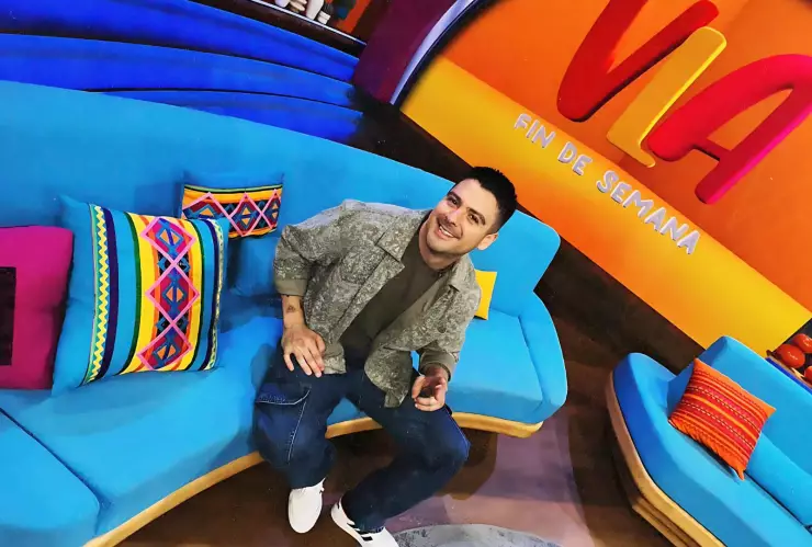 Conductor de televisión saludando y sentado en una sala color azul dentro de un foro de televisión con una pantalla gigante detrás