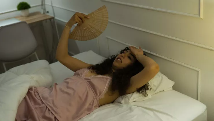 mujer aparentemente acalorada, est&aacute; recostada en su cama agitando un abanico 