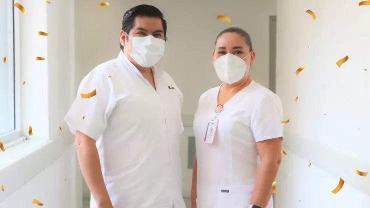 Un enfermo y una enfermera parados en un pasillo con uniforme color blanco. Los dos usan cubrebocas.