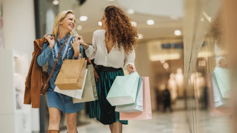 Dos mujeres caminan por el pasillo de un centro comercial cargadas de bolsas y riendo